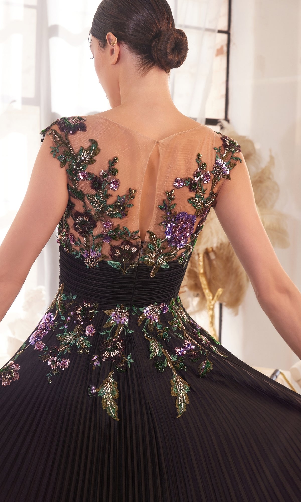 A woman is shown from behind wearing the Andrea & Leo A1355 Sequin Long Pleated Prom Dress, featuring sheer, embroidered floral details and sequin floral appliques on the back and shoulders.